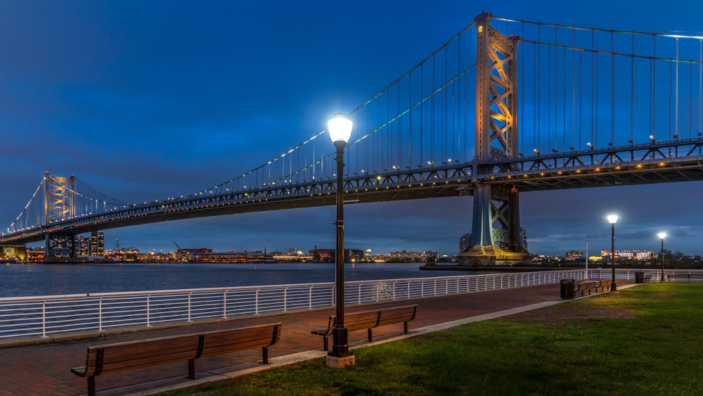 The Ben Franklin Bridge in Philadelphia, PA is seen at night, lit up against a dark blue sky. Old-fashioned streetlights and a patch of green grass in the foreground. Benches line the pathway along the water. 