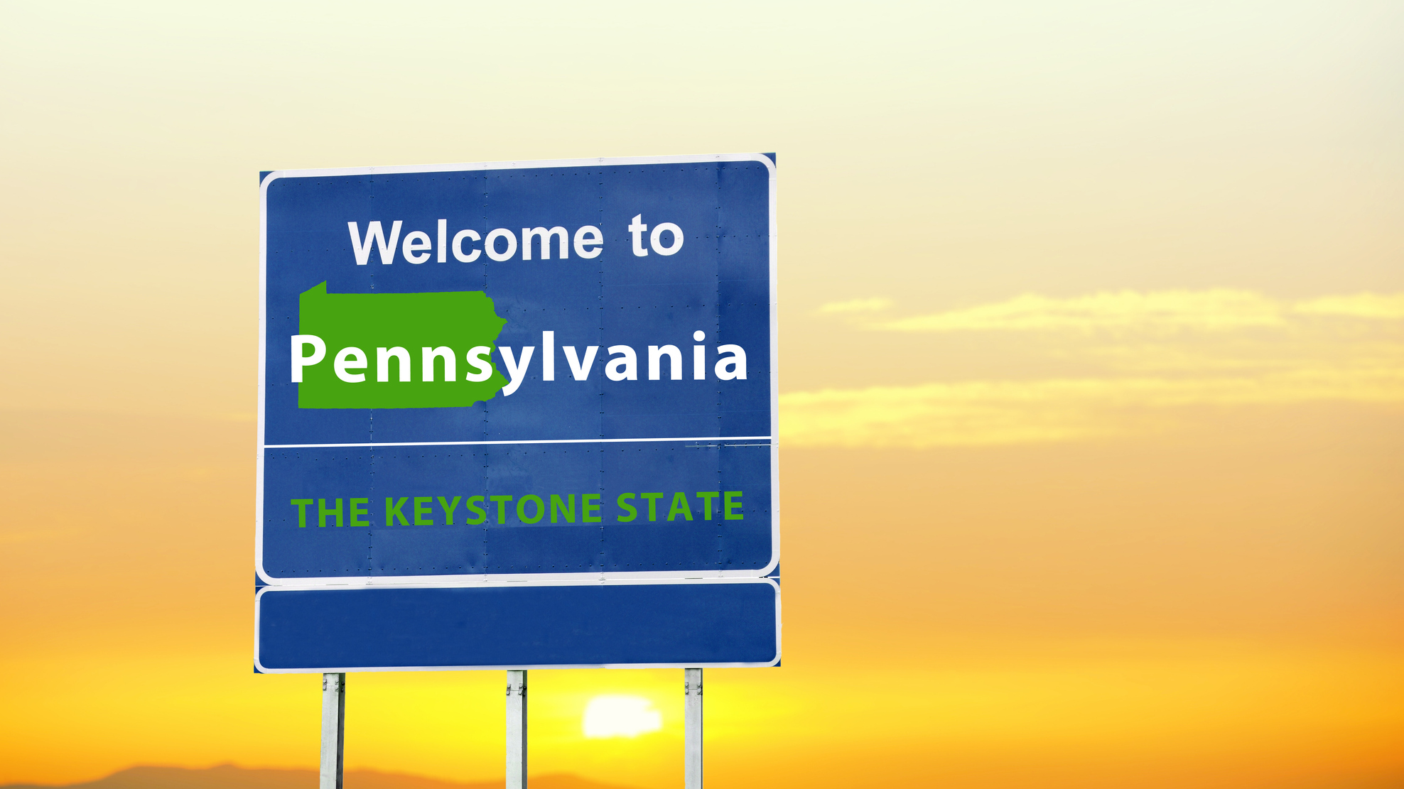 A "Welcome to Pennsylvania" road sign is seen against an orange sunset sky. the sign is blue and has a green outline of the state of pennsylvania. 